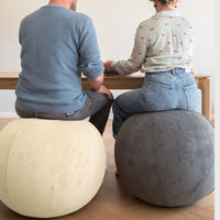 Two round floor sitting balls one cream and one dark gray corduroy, with a man and a woman sitting on them, viewed from behind in a bright room.