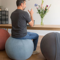 Man sitting on a balance ball in a home office setting with a desk and flowers.