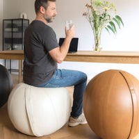 Man sitting on a white leather ball chair holding a glass of water in a home office setting.