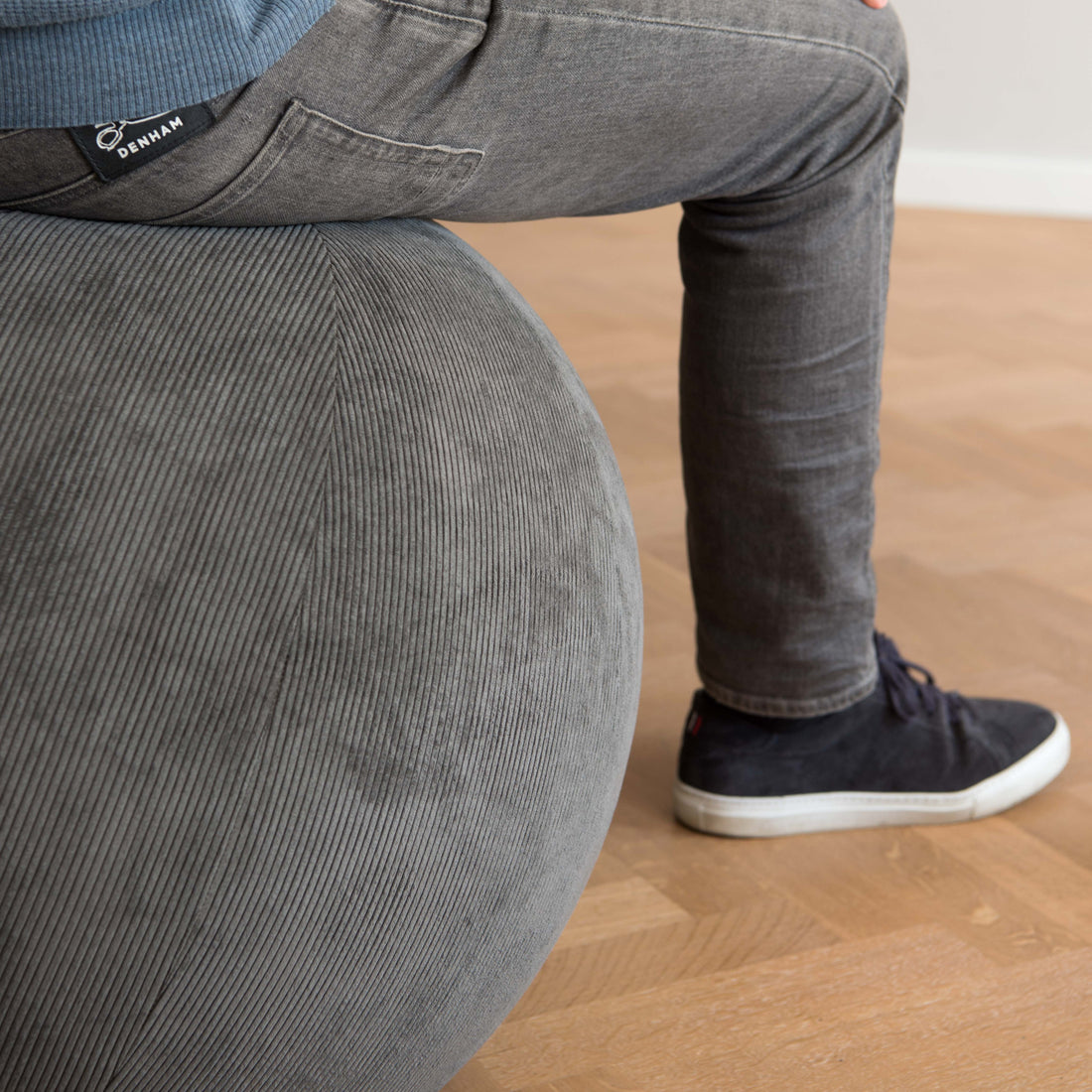 A close-up of a person sitting on a large gray corduroy exercise ball in an indoor room with a light wood floor. The person is wearing gray jeans, a blue ribbed top, and dark navy sneakers.