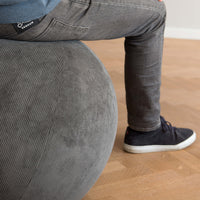 A close-up of a person sitting on a large gray corduroy exercise ball in an indoor room with a light wood floor. The person is wearing gray jeans, a blue ribbed top, and dark navy sneakers.
